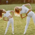 A mother and daughter in activewear stretching on a sunny day in a grassy field.
